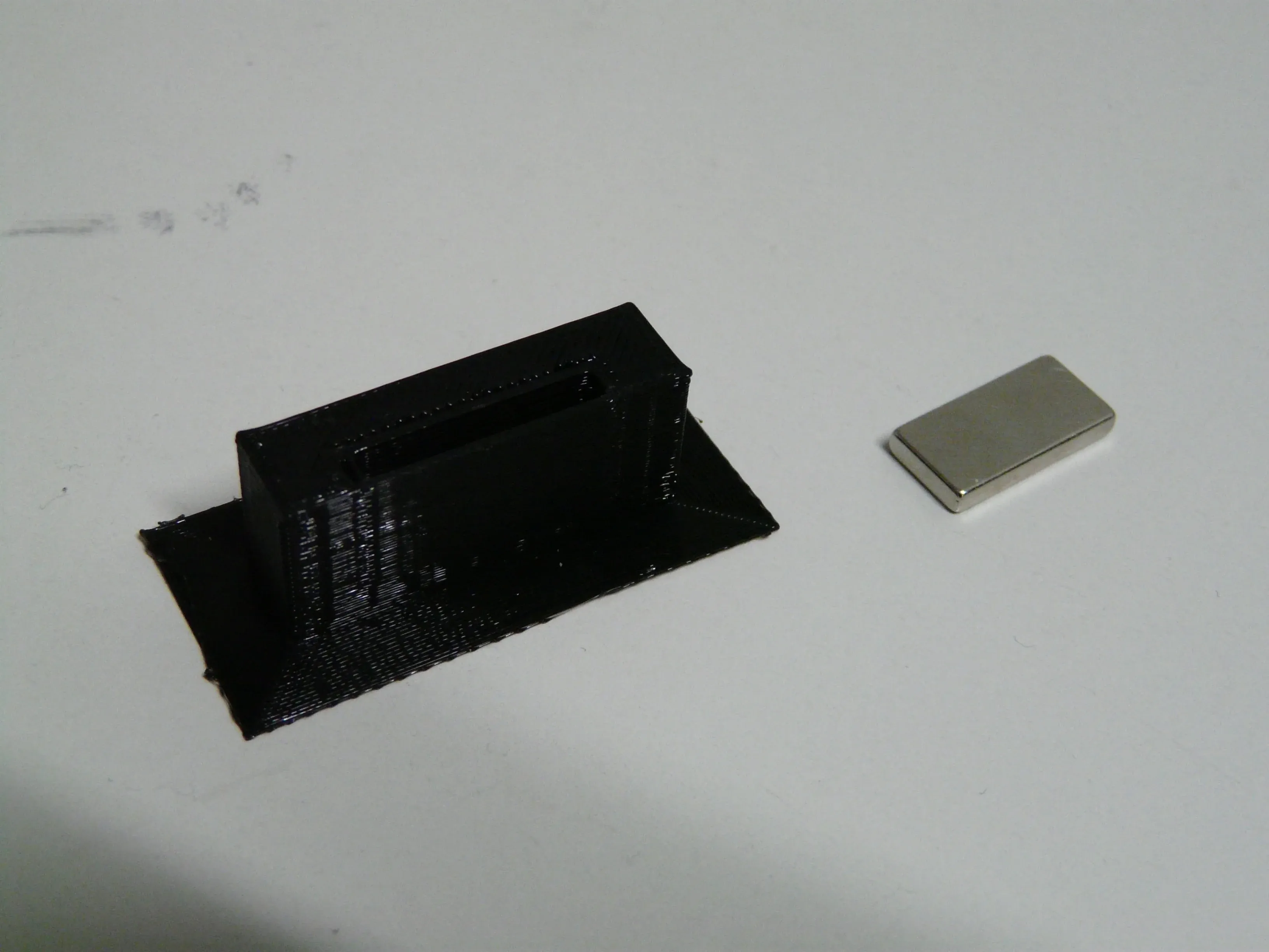 Musta 3D-tulostettu ovenpidike ja erillinen suorakulmainen magneetti.